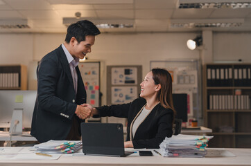 Two happy asian businesspeople shaking hands at an office desk in the evening, celebrating their successful deal and showcasing teamwork and collaboration in a professional environment