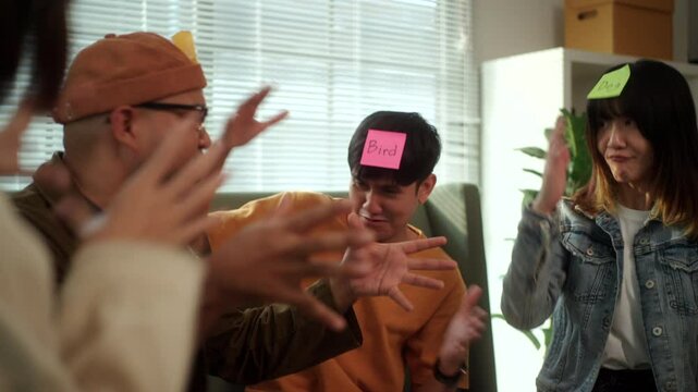 A group of young Asian friends is playing a fun charades game, guessing animal names at a lively home party on the couch. Laughter fills the room as they try to guess each other's clues.
