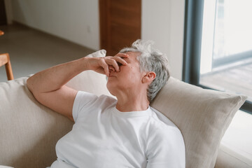 A senior woman with white hair sits on a sofa in the living room and holds her head in her hands, she has a headache and feels unwell. She drinks water and covers her face with her hand. Medical care.