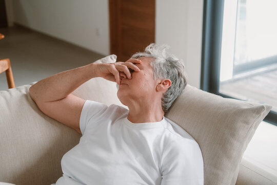 A senior woman with white hair sits on a sofa in the living room and holds her head in her hands, she has a headache and feels unwell. She drinks water and covers her face with her hand. Medical care.