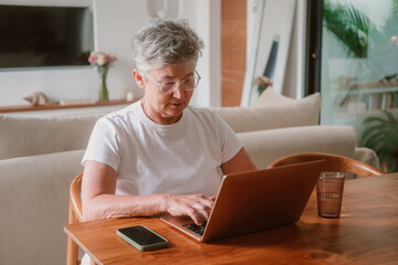 Portrait of a senior woman with gray hair wearing glasses working on a laptop at home in the living room. She is typing on a computer and working remotely from home. Remote work skills for retirees.