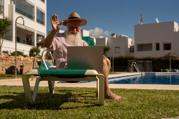 Smiling retired man making a video call on his laptop while sitting by the pool at a Spanish resort, enjoying relaxed retirement life and digital communication on vacation