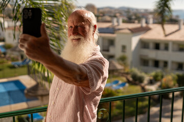 Happy senior man with long white beard taking selfie with smartphone on balcony, enjoying sunny vacation in tropical resort