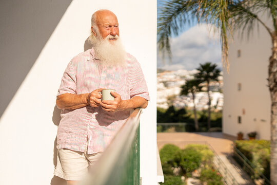 Smiling elderly man enjoying morning coffee in sunlight while standing on balcony with palm trees in the background - Powered by Adobe