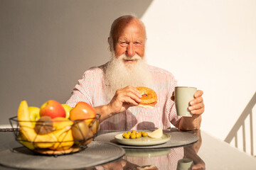 Smiling elderly man holding a coffee cup and bagel during breakfast, enjoying healthy food and morning sunshine at a modern table with fresh fruits
