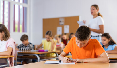Diligent elementary school student tween boy studying with classmates, making notes of teacher lecture