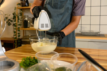 Man in black gloves mixing cream in a clear glass bowl with an electric hand mixer