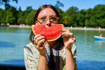 Young woman holding slice of watermelon in front of face in park