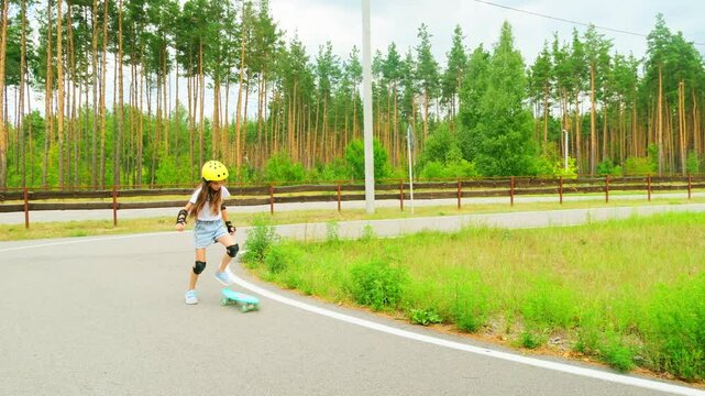 Girl in helmet and knee pads learning to skateboard. Child skateboarding in forest park.
