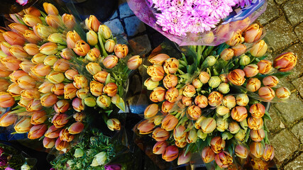 Top view of orange and yellow tulip bouquets wrapped in plastic at a flower market. Fresh spring blooms displayed on rustic stone pavement, radiating warmth and vibrant seasonal beauty.