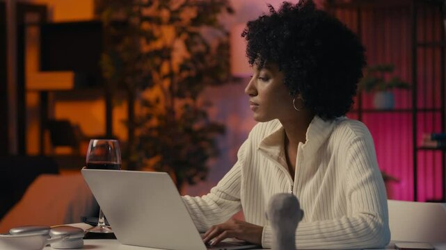 Young African American woman drinking wine and working on laptop late at night
