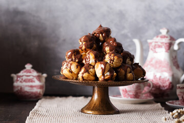 Profiterole cake, or cream puff, is a French choux pastry ball filled with whipped cream, custard, and chocolate sauce, served on a gold plate with cup of tea and teapot.