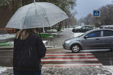 Woman under an umbrella in a heavy snowfall crosses the road at a crossroads. Bad weather, climate change, mud and puddles. Public utilities do not have time to clear the roads