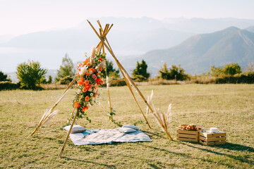 Tipi-style wedding arch stands next to wooden boxes on a green meadow in the mountains