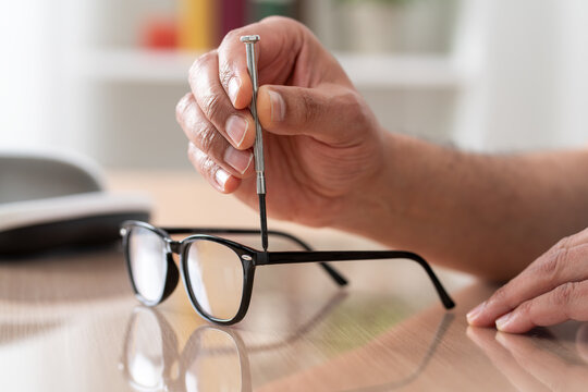 Close up of man hands tightening screw on glasses with screwdriver on a desk at home.Male optician repairing and fixing eye glasses with screwdriver.