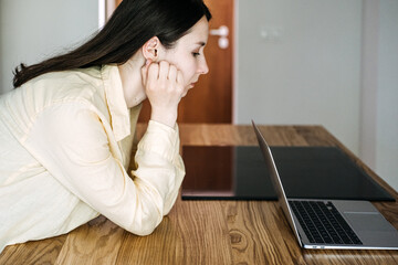 Young Asian woman sitting at a wooden table, staring at a laptop screen with a serious expression. Job insecurity, AI disruption, remote work stress, automation impact
