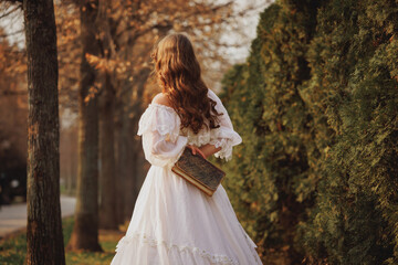 Fototapeta premium Girl holds vintage book behind her back while walking through autumn park in soft golden light, carrying echoes of past. Concept of vintage, comparison of eras, fantasy. Book or film cover.