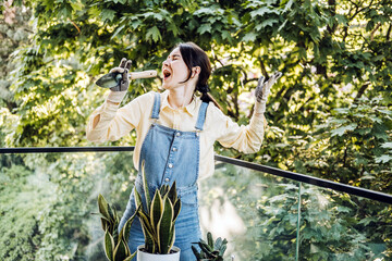 Young Asian woman playfully singing into a garden trowel while caring for balcony plants. Plant...