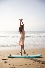 She stands barefoot in pointe shoes on a surfboard, pink dress floating in the breeze a lighthearted moment filled with beach vibes, freedom, and graceful joy under the sun. 