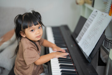 Young Asian girl enjoy plays piano at home. Little kid prcatices music instrument in house. Child interested in musical, pressing on keyboard and looking at camera.