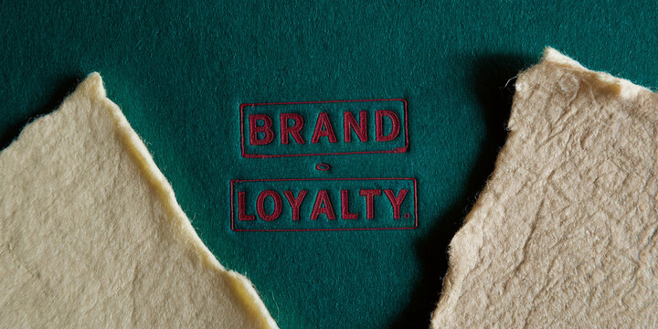 Green textured background with beige paper scraps, featuring red embossed words "Brand Loyalty", showcasing a concept of brand dedication and customer commitment