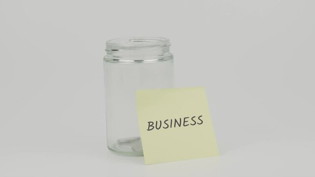 Business text on a yellow note stuck to a jar of coins, with a hand inserting a coin. The image symbolizes financial growth, investment, and business management, set against a white background