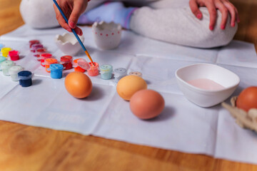 Child is dipping a brush in orange gouache, preparing to paint easter eggs on a white sheet on a wooden table, with other eggs and a bowl nearby