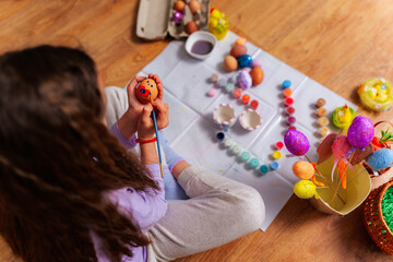 Young girl sitting on the floor painting an easter egg with a face on it, surrounded by colorful...