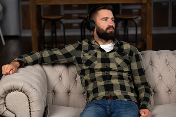 Fototapeta premium A bearded man sitting on a sofa, wearing headphones, lost in thought. Deep thinking, relaxation, personal reflection, or moments of contemplation.