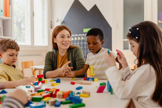 Smiling teacher interacts with diverse group of young children playing with colorful blocks in a bright, cheerful classroom setting.