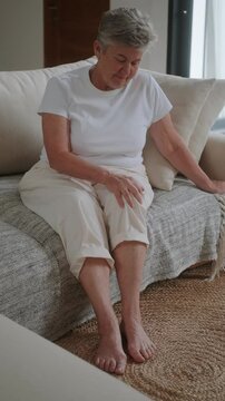 A senior woman with white hair sits on sofa in living room and rubs her knee with her hand because she feels pain. Knee and joint pain in people of retirement age. Arthritis and joint wear in elderly.