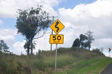 A yellow road sign indicating winding road ahead with a speed limit of 50 km/h, set against a rural...