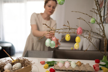 A woman enthusiastically decorates a lovely branch with delightful pastel Easter eggs for a joyful...