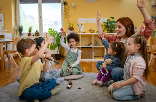 Happy kindergarten teacher and a diverse group of children raising their hands together during music time in a bright classroom. - Powered by Adobe
