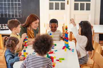 Diverse preschool children play with colorful building blocks at a classroom table, guided by a...