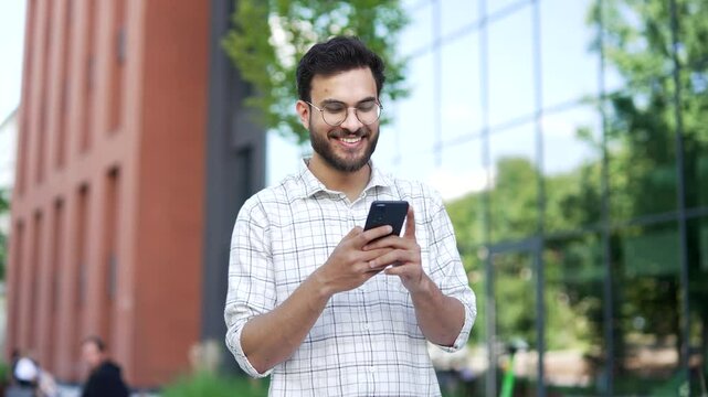 Smiling handsome man is using a mobile phone while walking on the street near an modern building. Happy bearded male chatting online, writing reading messages, browsing the web or social media