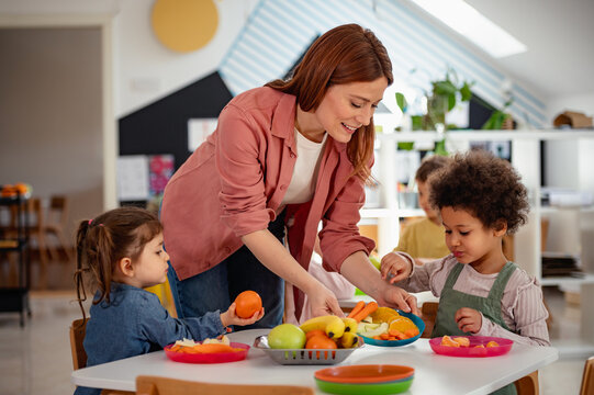 A diverse group of preschool children sits around a table eating healthy snacks while a teacher engages with them, promoting nutrition, inclusion, and social interaction in early education.