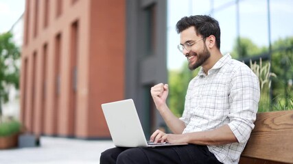 Satisfied happy handsome man received good news on laptop while sitting on a bench on the street near office building. Smiling glad businessman reads a positive good message on computer, rejoices