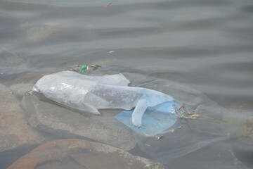 Plastic waste floats on the surface of calm water near rocky shoreline during overcast day