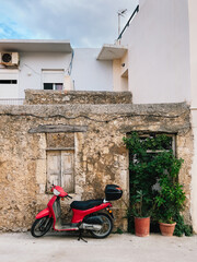 Red scooter parked near an old stone house with wooden shutters and potted plants in typical Greek village, showcasing charm of Mediterranean architecture and lifestyle. Red scooter, stone house © Viktar Vysotski