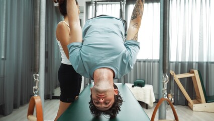 Healthy client practicing inverted leg raise on Pilates Cadillac under instructor guidance. Man hanging back stretch on the cadillac for improving core strength, and upper body stability. Habituate.