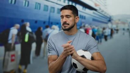 Man holding soccer ball at seaside port with cruise ship and crowd in background, capturing a moment of anticipation and casual outdoor leisure with sports theme.