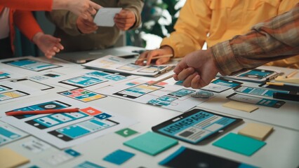 Fototapeta premium A diverse team engages in a productive brainstorming session, developing mobile app wireframes and design concepts on a bright, organized workspace table. SACTR
