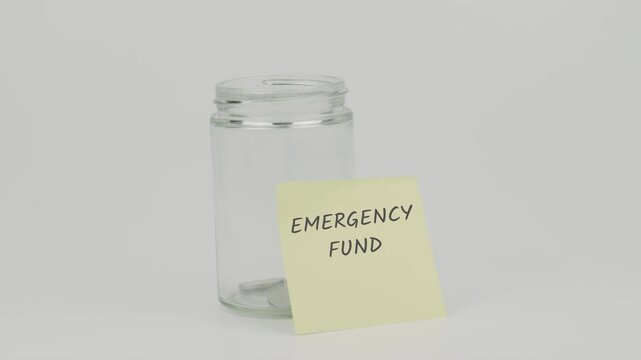 Emergency Fund text on a yellow note stuck to a jar of coins, with a hand inserting a coin. The image represents saving for unexpected expenses and financial preparedness, set against a white