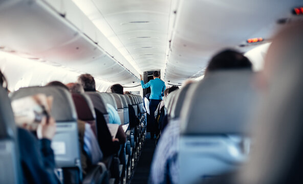 Fototapeta Interior of airplane with passengers on seats and stewardess in uniform walking the aisle, serving people. Commercial economy flight service concept