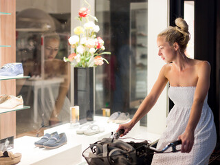 Casual young lady on bicycle wearing white summer dress window shopping in front of sinfully expensive boutique store display window. Customer woman in shopping street looking at window an night.