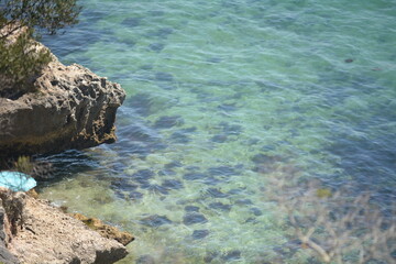 Clear turquoise water laps against rocky coastline under bright sunlight near a secluded beach in summer