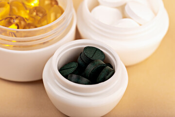Green spirulina tablets in white cosmetic container with yellow softgel capsules and white pills blurred in background. Concept of health and dietary supplements. Closeup of spirulina tablets in jar