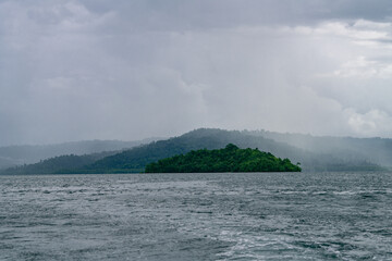 A small green island emerges through a tropical rainstorm surrounded by vast ocean and misty jungle hills