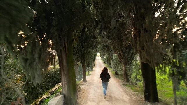 caucasian brunette young woman in leather jacket sunglasses blue jeans enjoying walking, running at the Italian cypress tree alley in Tuscany in sunny day